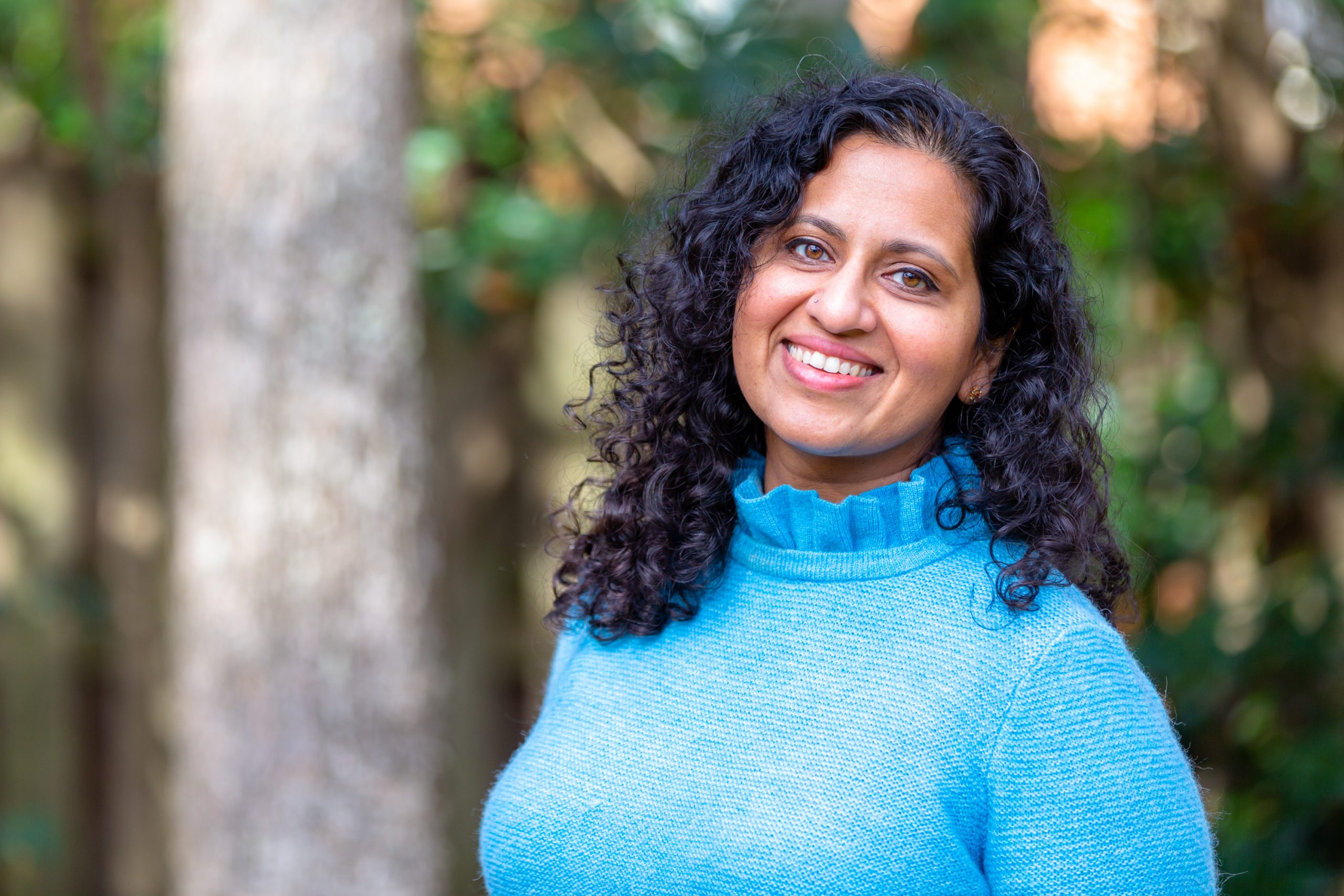 Picture of Aparna Polavarapu wearing a blue sweater, standing in front of trees